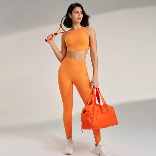 Woman in orange athletic wear holding a tennis racket and bag on a plain background